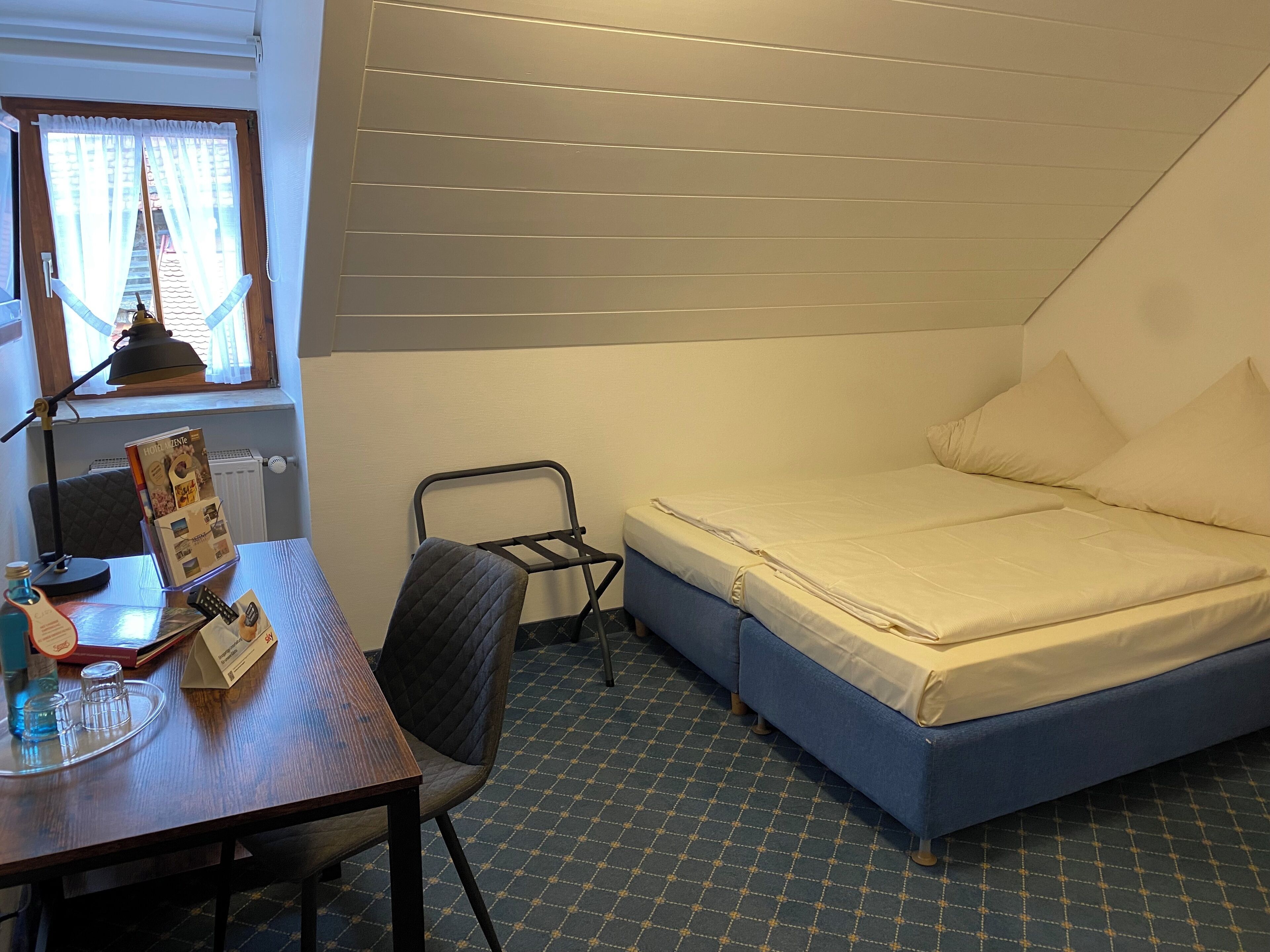 Een kleine, verzorgde slaapkamer met een enkel bed, een bureau met stoel, een radiator en een raam met gordijnen.
