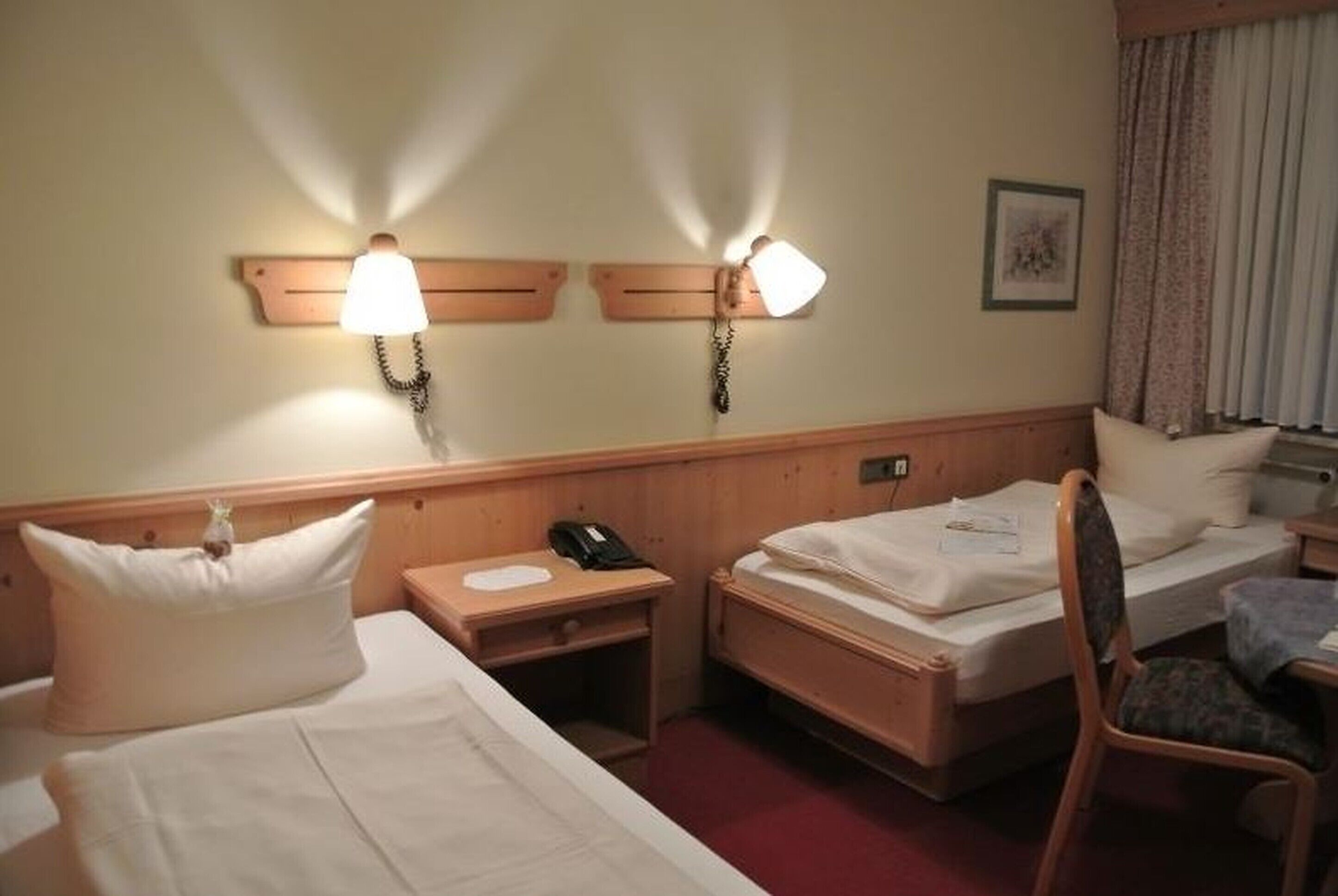 A hotel room with two single beds, a wooden headboard, a nightstand with a phone, a desk with a lamp, and a chair.