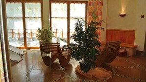 Una habitación con suelo de baldosas, un banco de madera y dos sillas. Hay plantas en macetas y una mesa con sillas.