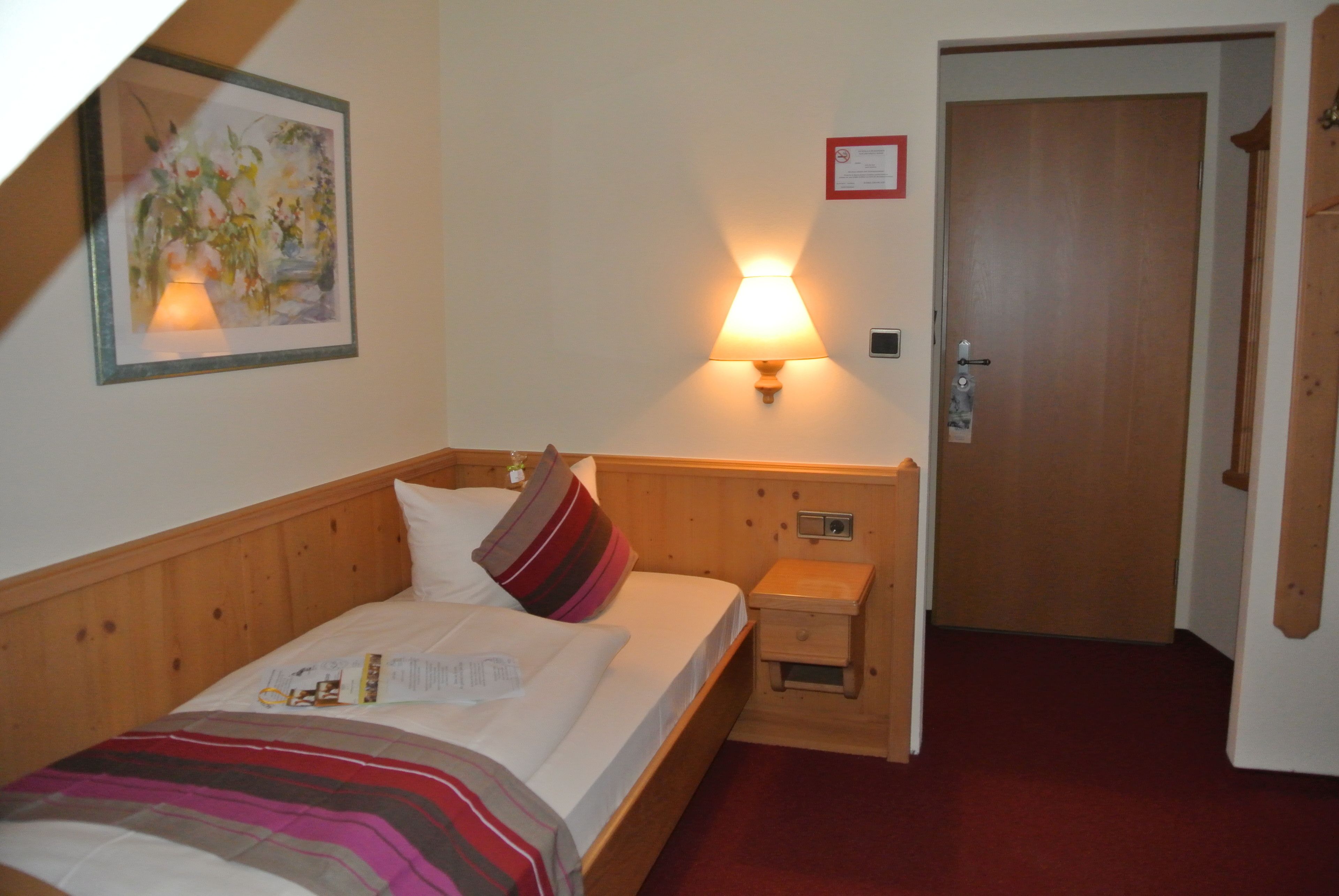 Ett hotellrum med en säng i trä, en sänglampa, en inramad tavla på väggen och en dörr med lås.