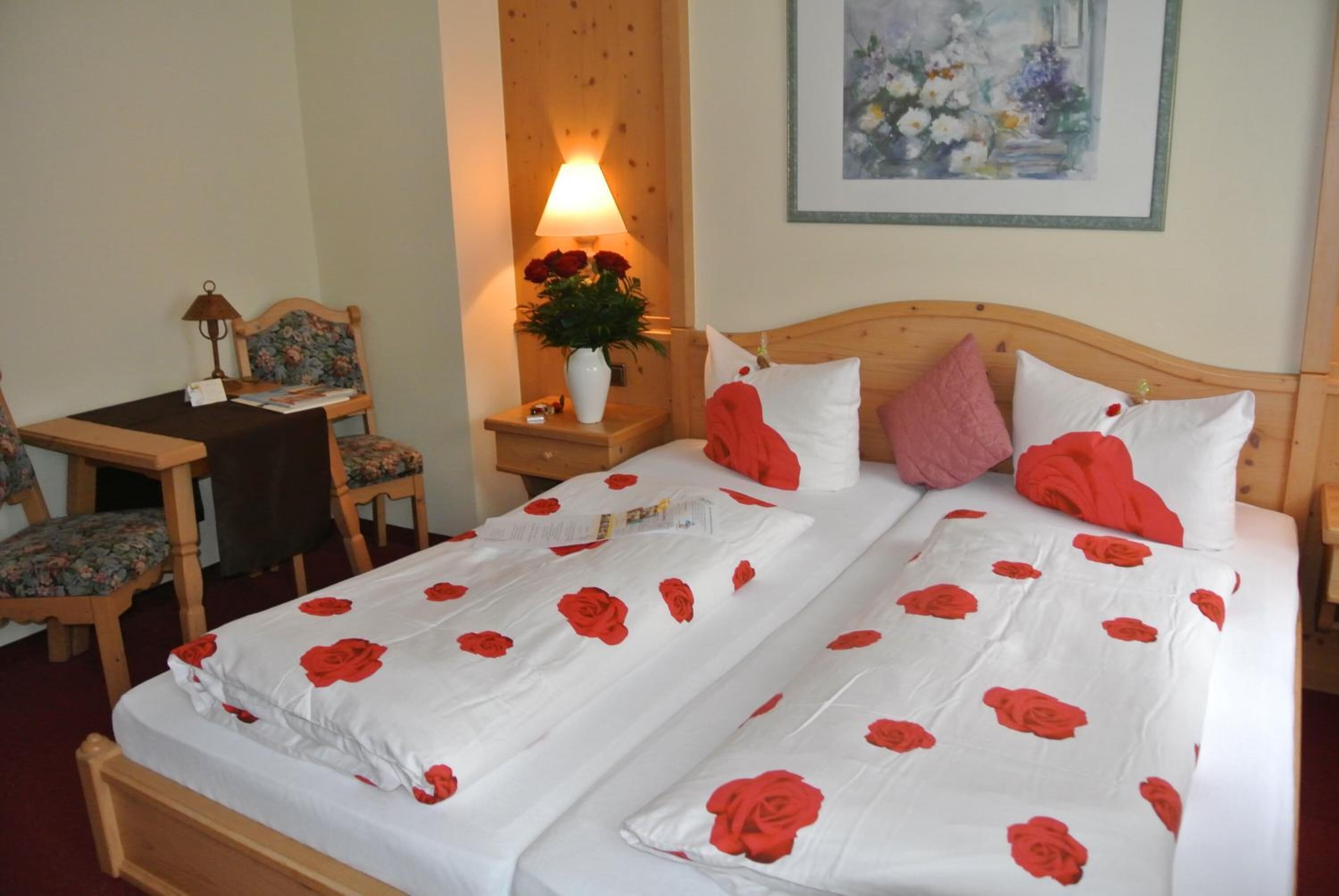 A double bed with white bedding and red rose patterns, a wooden nightstand with a lamp and vase of flowers, a wooden chair, and a framed painting on the wall.