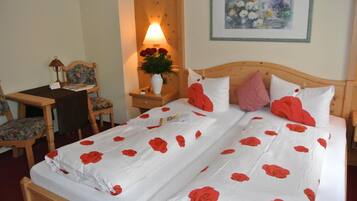 A double bed with white bedding and red rose patterns, a wooden nightstand with a lamp and vase of flowers, a wooden chair, and a framed painting on the wall.