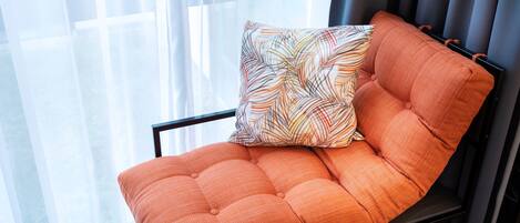 Una silla acolchada con un cojín estampado, ubicada cerca de una ventana con cortinas traslúcidas.