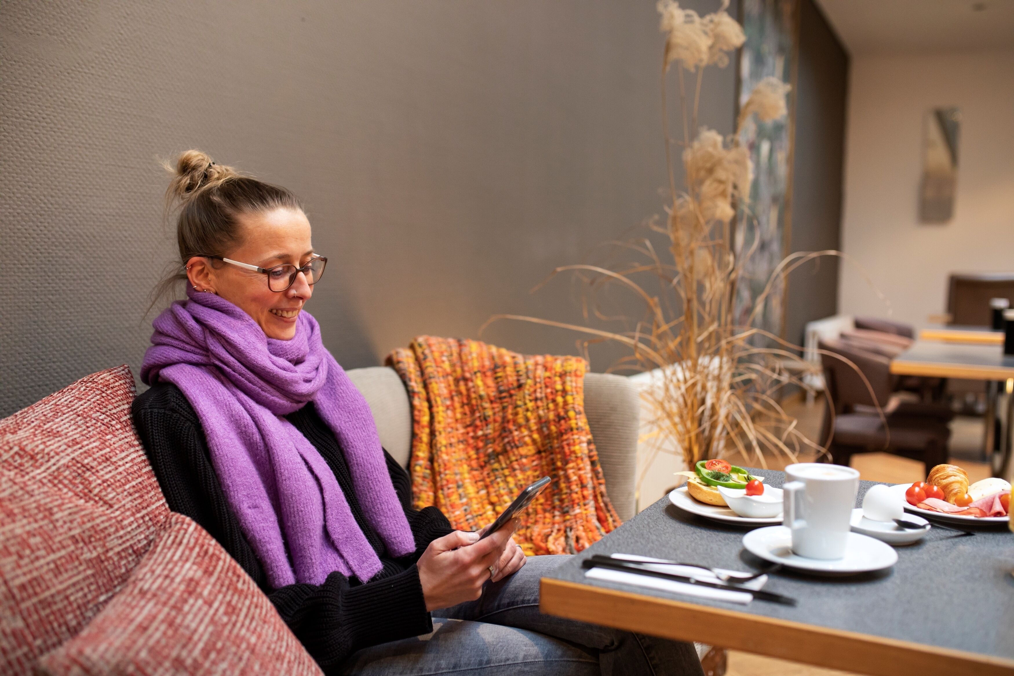 Una persona sentada en un sofá usando un teléfono inteligente, con una mesa frente a ella que tiene un plato de comida, una taza y un croissant.