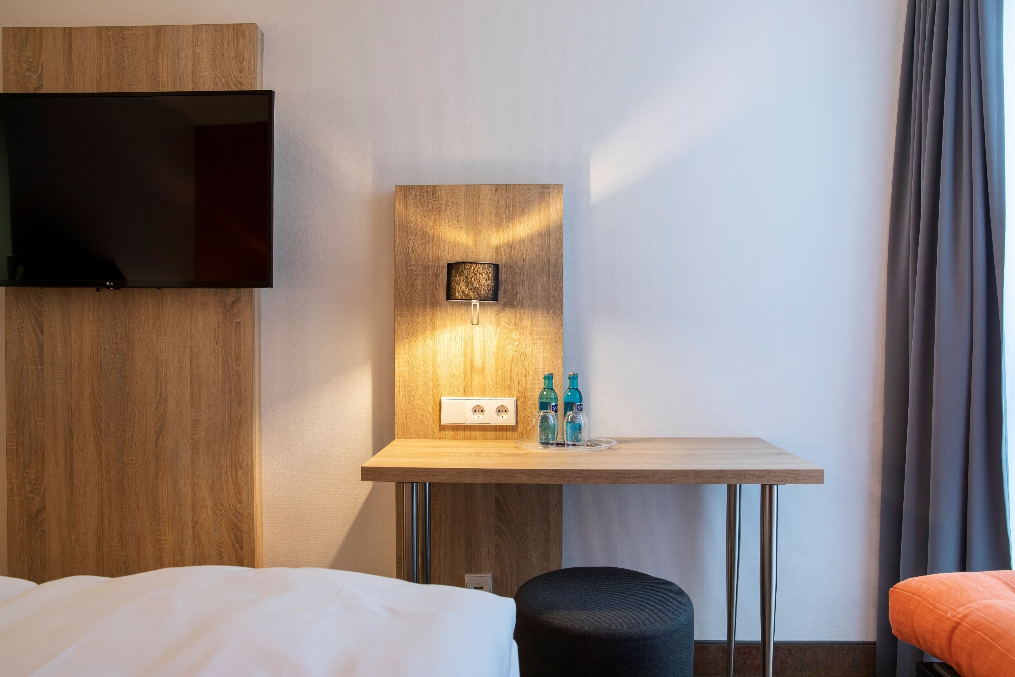 A hotel room with a wooden desk, a flat-screen TV mounted on the wall, a small table with two bottles, and a bed with white bedding.