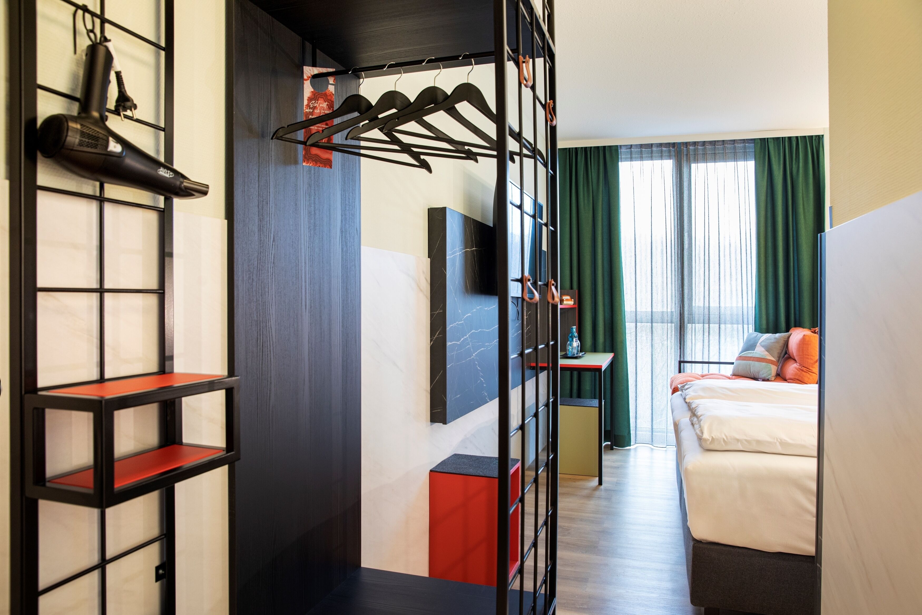 Ein Hotelzimmer mit Bett, Wandfernseher, Schuhablage und Regal mit roter Grundplatte.