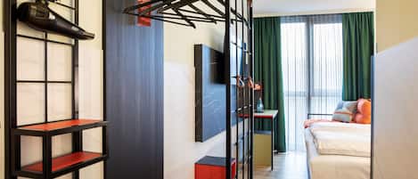 A hotel room with a bed, a TV mounted on the wall, a shoe rack, and a shelf with a red base.