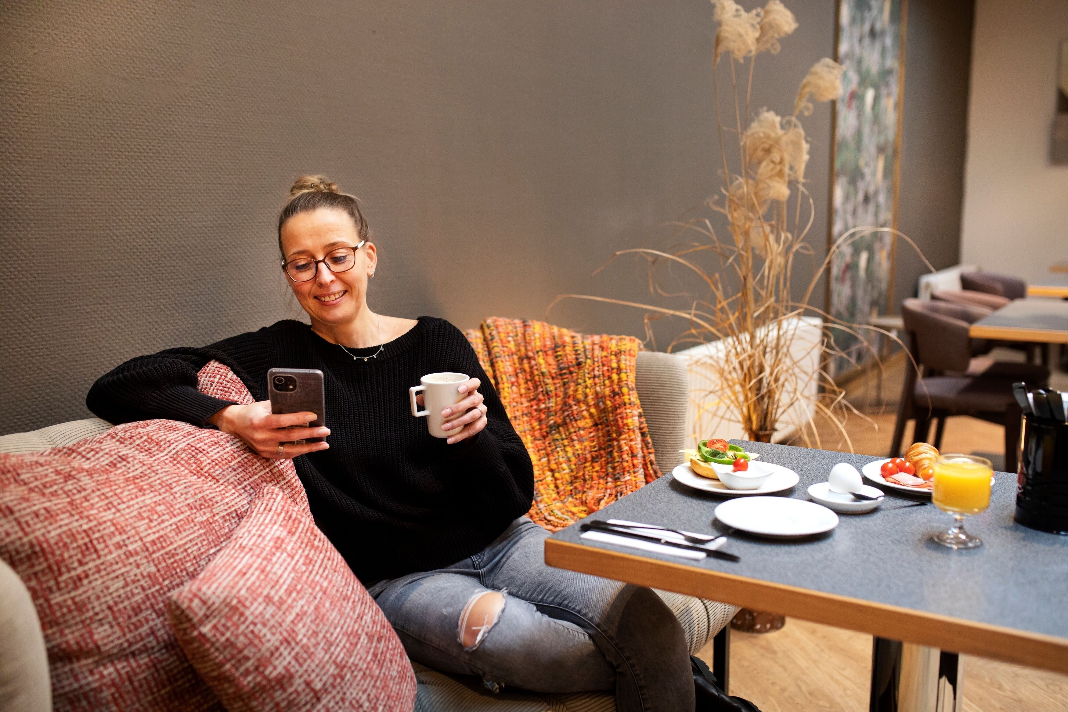 Una persona sentada en un sofá con un teléfono inteligente y una taza, una mesa con alimentos para el desayuno, y un florero con plantas secas.