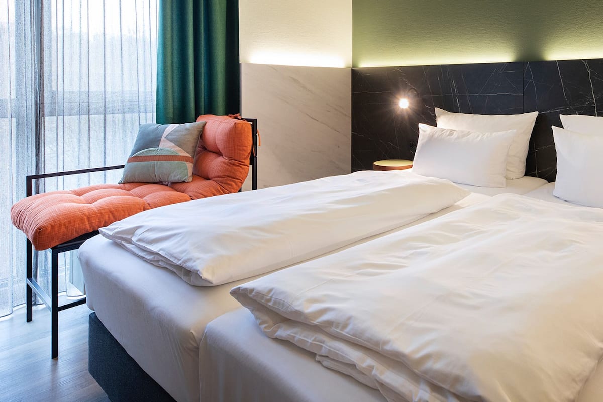 A hotel room with a large bed, two bedside tables, a chair with cushions, and a view through a window with curtains.