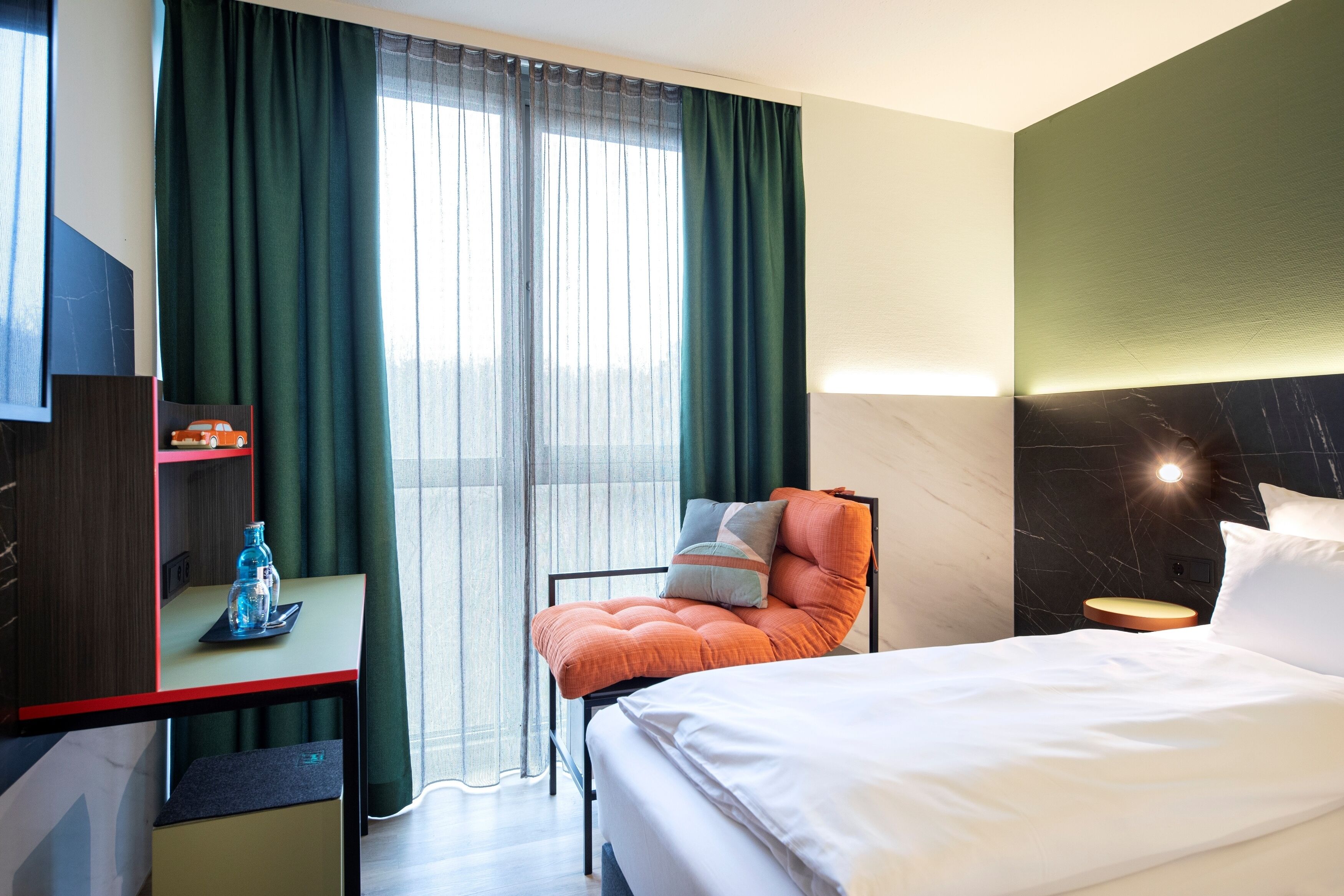 Una camera d'albergo con un letto, una sedia, una scrivania con una bottiglia d'acqua e una TV su un mobile.