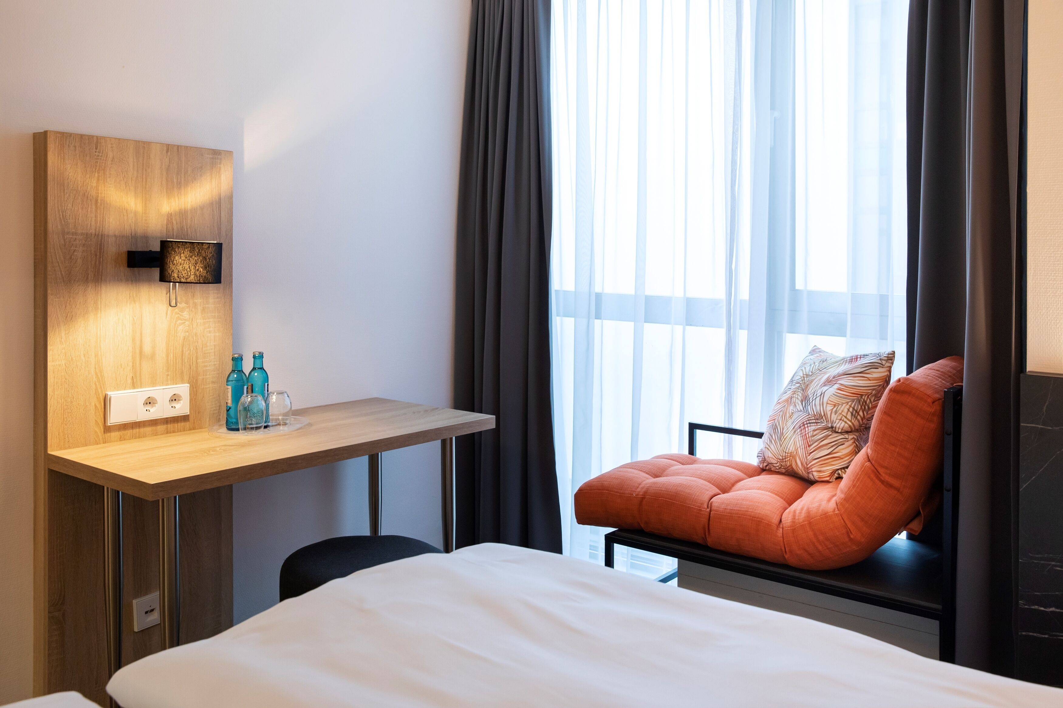 Une chambre d’hôtel avec un lit, un bureau avec une lampe, une chaise et une fenêtre avec des rideaux.