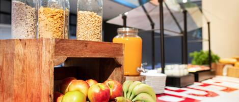 A breakfast buffet with a wooden crate containing apples and bananas, a glass jar of orange juice, and containers of cereal and grains.