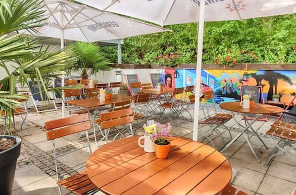 Um café ao ar livre com mesas e cadeiras de madeira, guarda-sóis e um mural na parede.