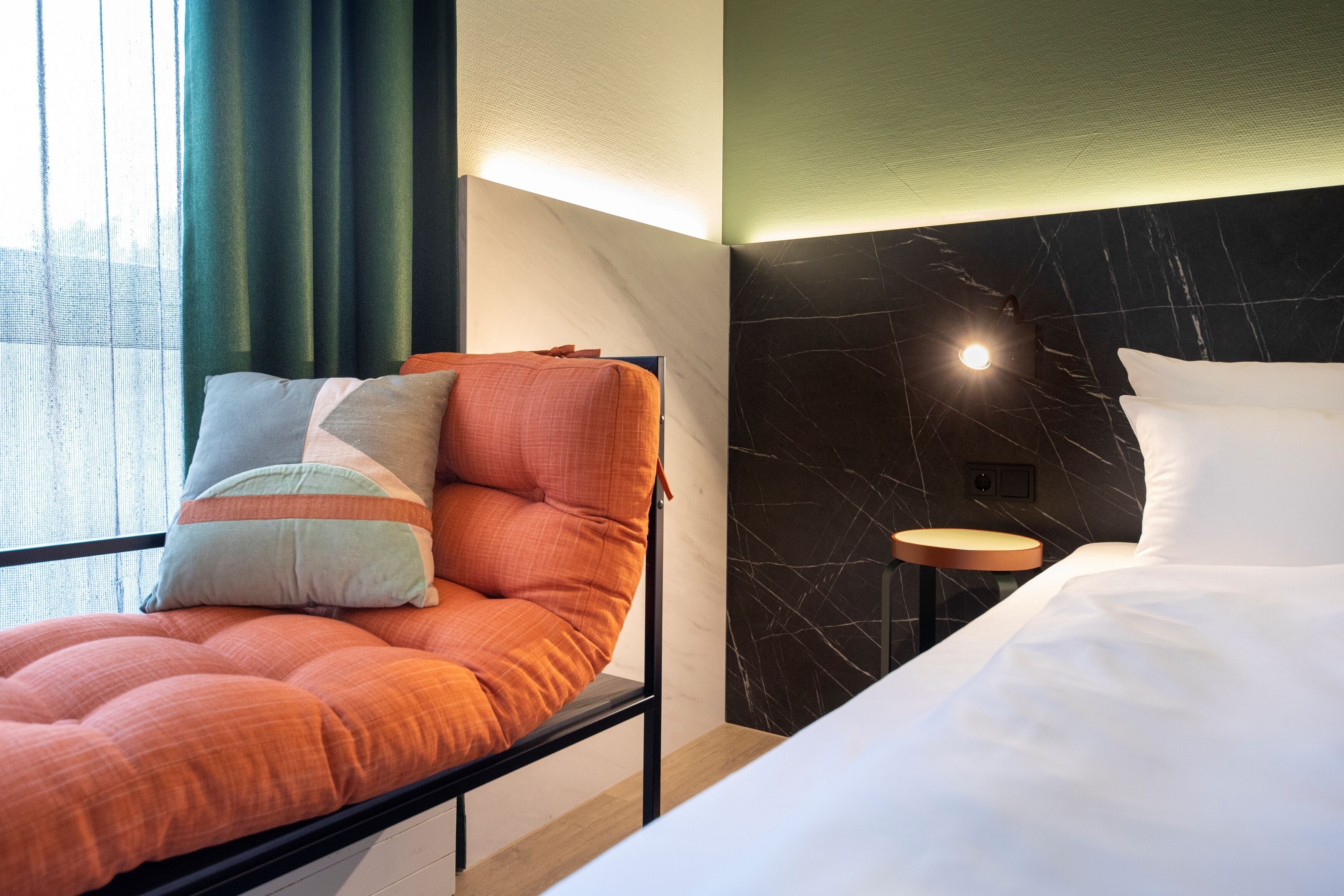 Habitación de hotel moderna con una cama, un rincón de descanso con cojines, una mesita y vistas a través de una ventana con cortinas.