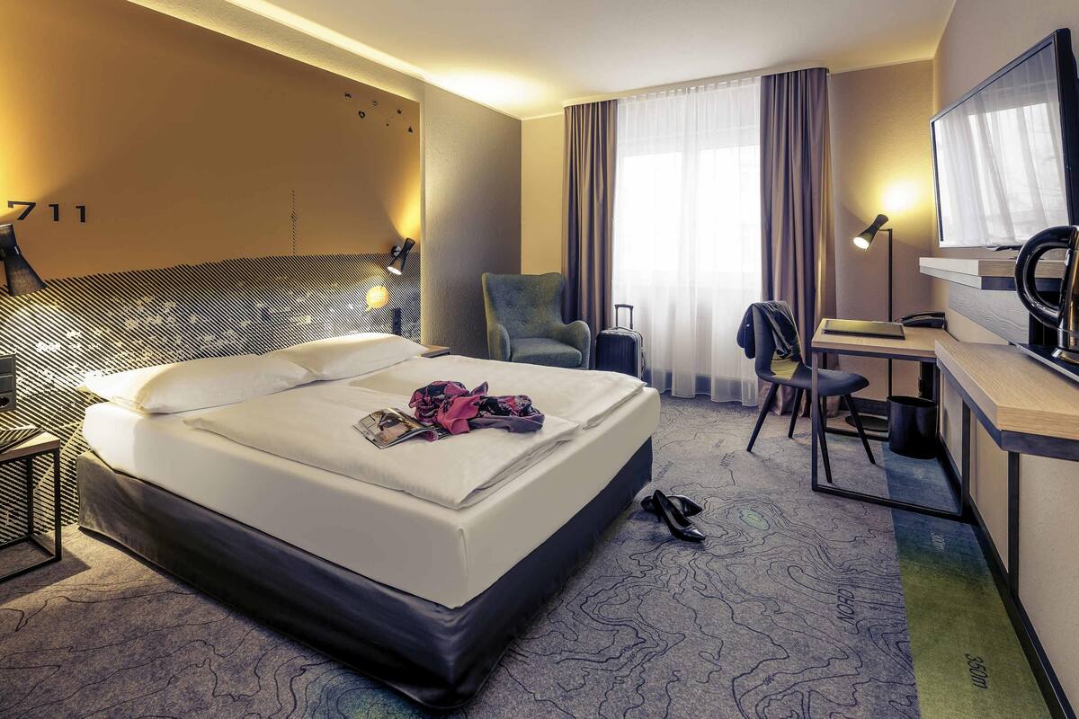 A hotel room with a bed, desk, chair, and a large window with curtains.