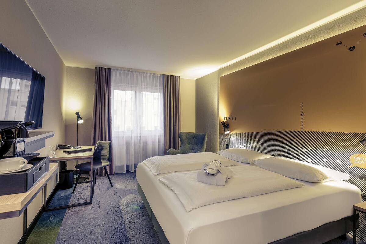 A hotel room with a large bed, a desk with a chair, a desk lamp, and a window with curtains.
