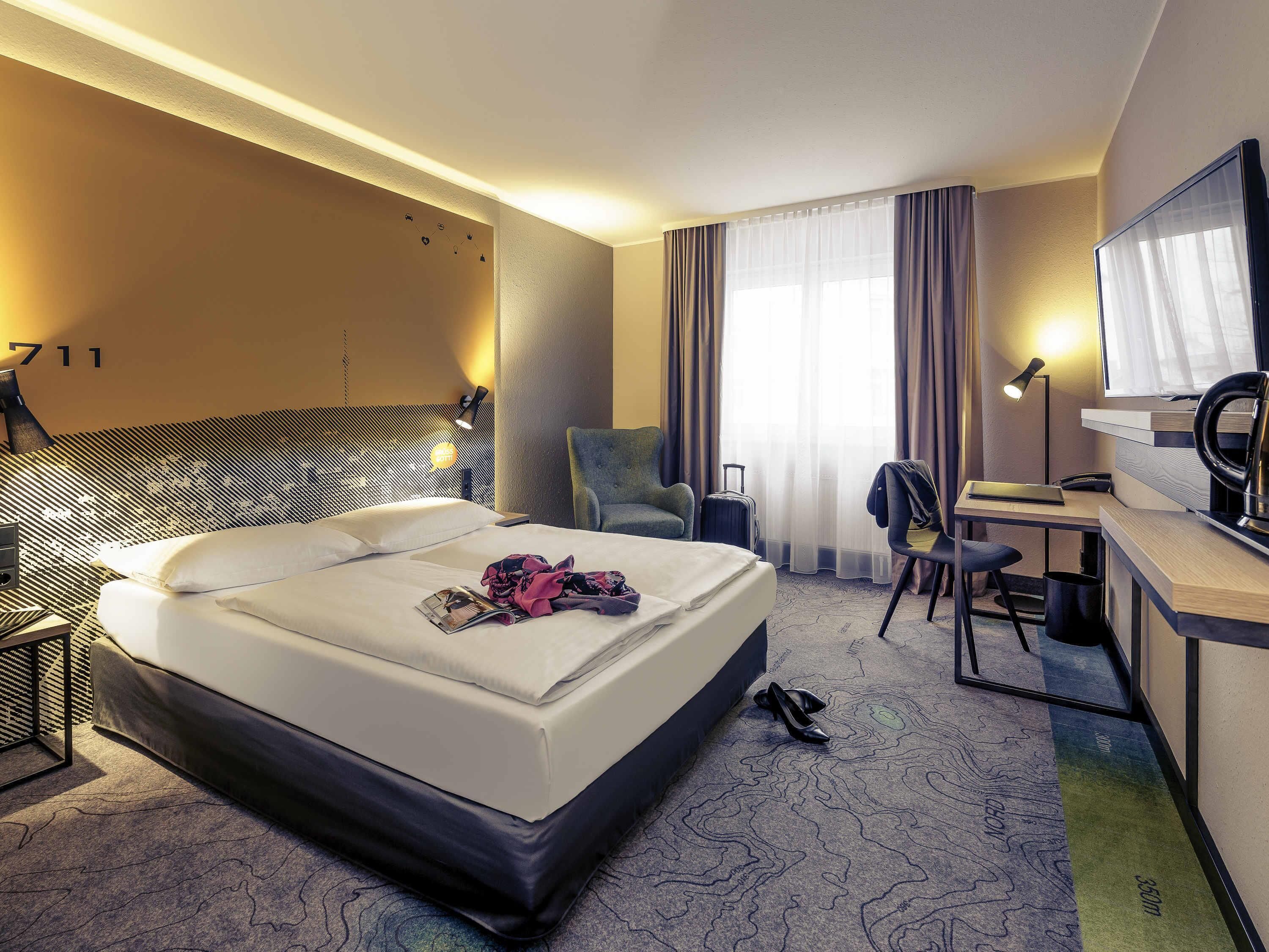 A hotel room with a bed, desk, chair, and a large window with curtains.