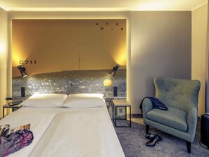 Een hotelkamer met een groot bed, een hoofdbord met een patroon, twee bedlampjes, een stoel en een koffer.