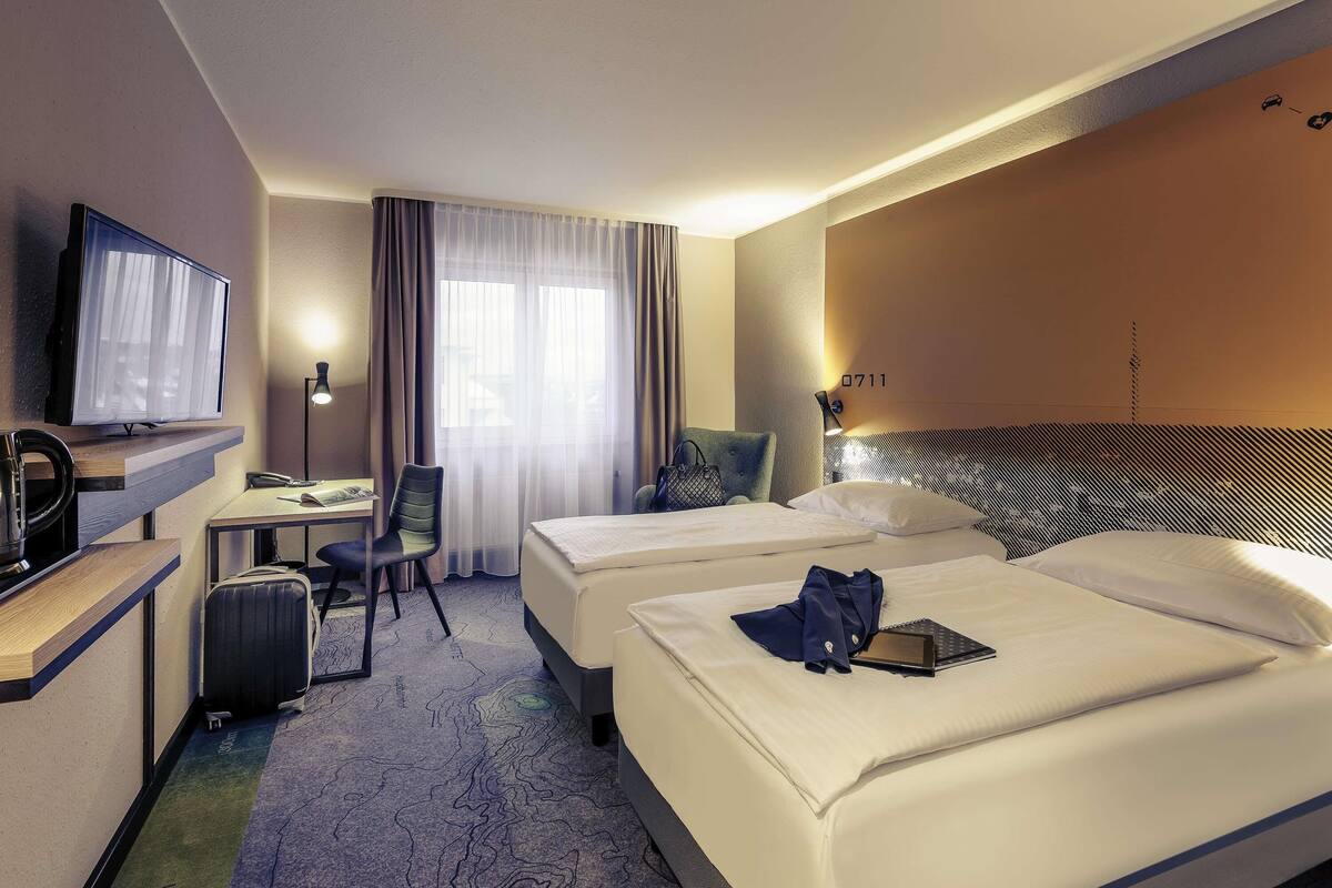 A hotel room with two beds, a desk with a chair, a television, and a window with curtains.