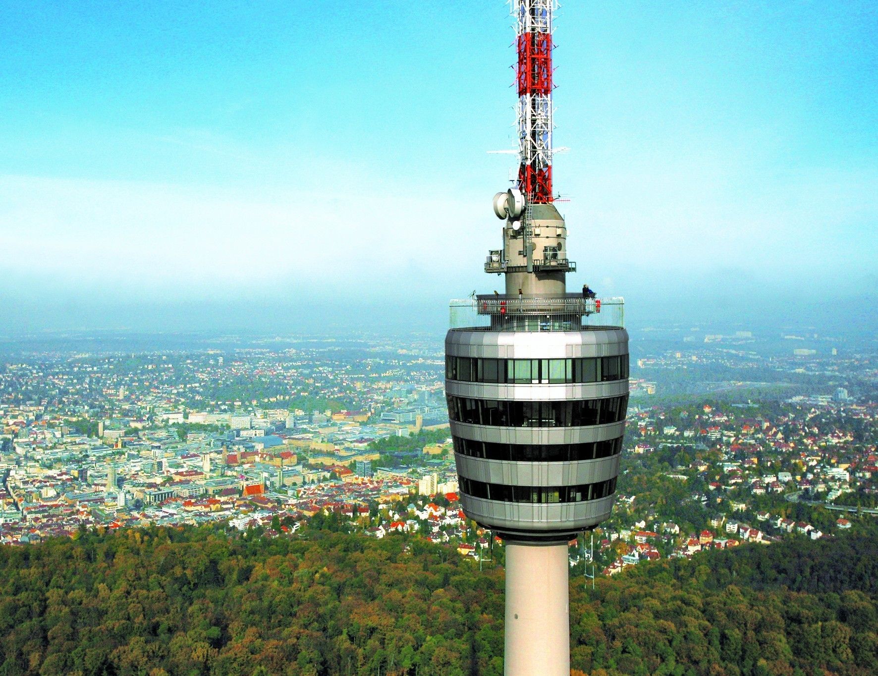 Una torre con un mirador circular, rodeada por un área boscosa y un paisaje urbano al fondo.