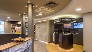 A hotel reception area with a reception desk, a wall with a pattern, and a display screen. - Mercure Hotel Stuttgart Zuffenhausen (Stuttgart)