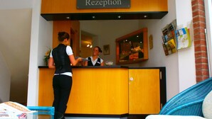A reception area with a wooden reception desk, a receptionist, and a guest standing. - Hotel Am Braunen Hirsch (Celle)