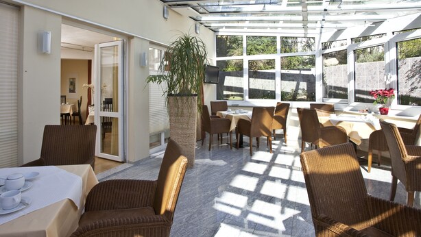 A sunlit patio with wicker furniture, a large potted plant, and a glass-enclosed dining area. - Hotel Am Braunen Hirsch (Celle)