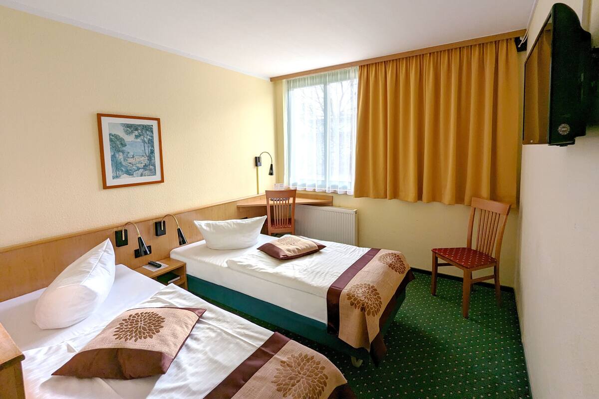 A hotel room with two beds, a desk, a chair, a window with curtains, a radiator, and a picture on the wall.