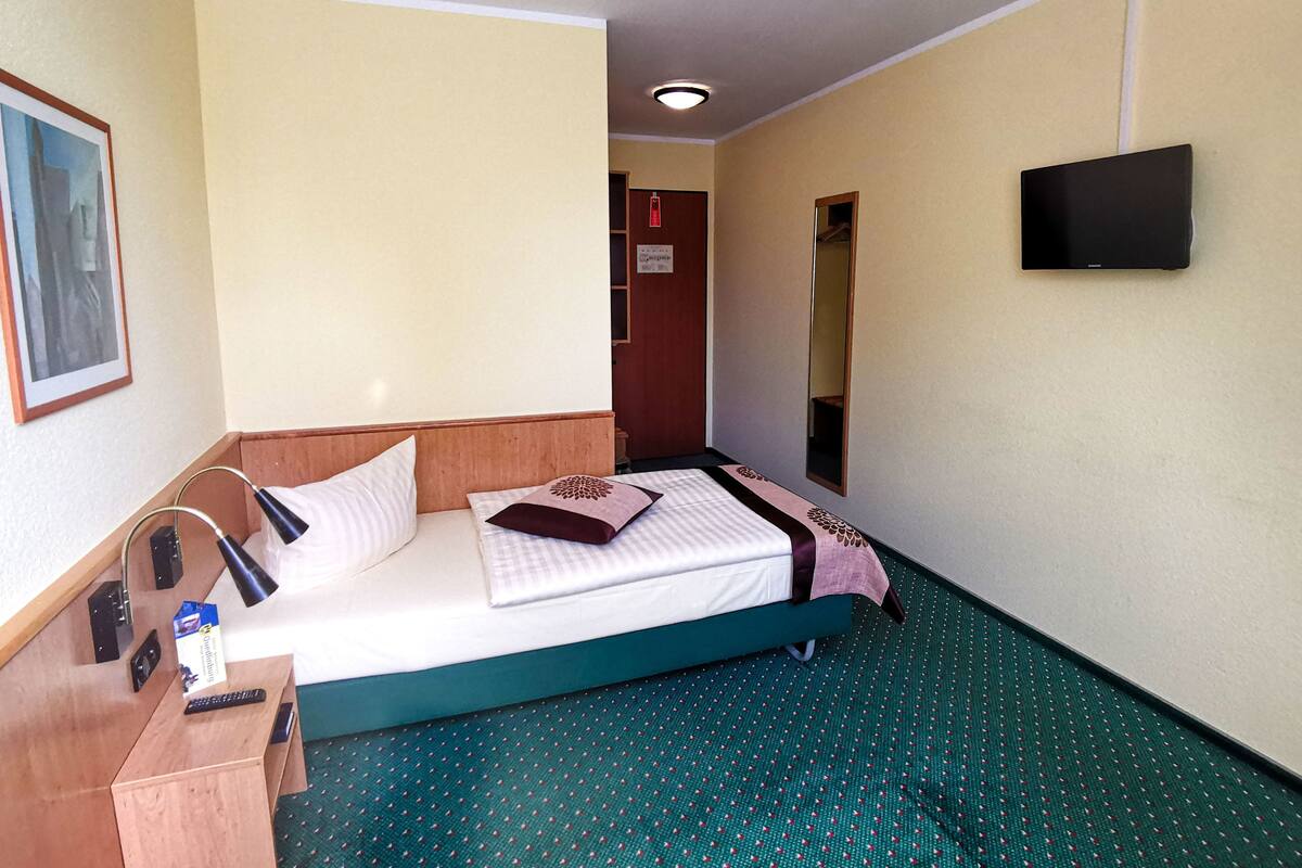 A hotel room with a bed, a desk with a lamp, a TV mounted on the wall, and a door leading to another room.