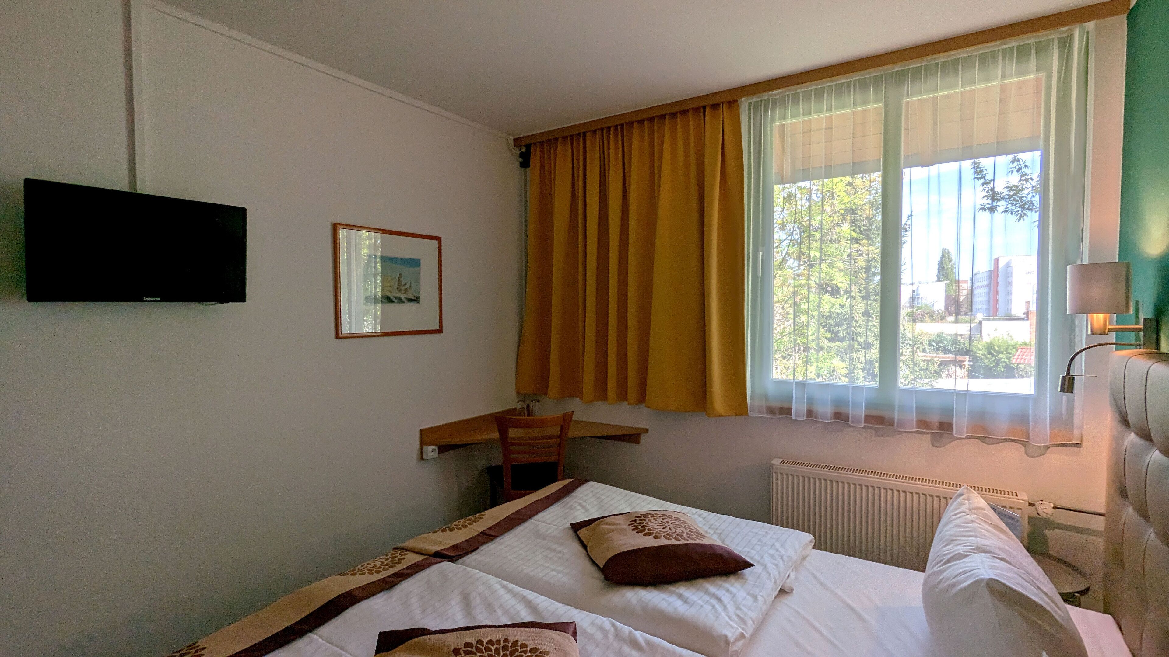 Una camera d'albergo con un letto, una scrivania, una televisione, una finestra con tende e un termosifone.