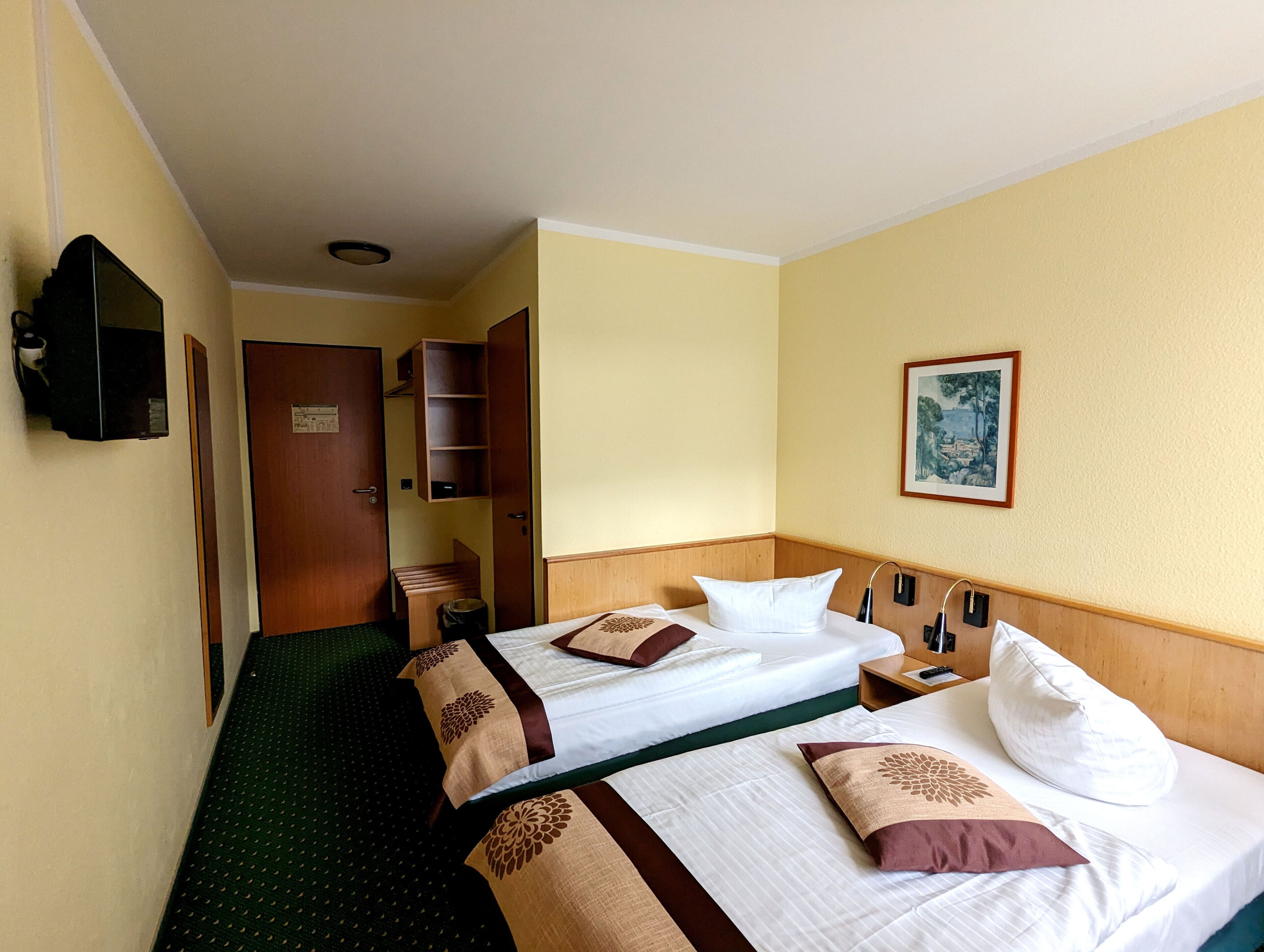 A hotel room with two beds, a wooden headboard, a small wooden cabinet, a trash bin, a framed picture on the wall, and a television mounted on the wall.