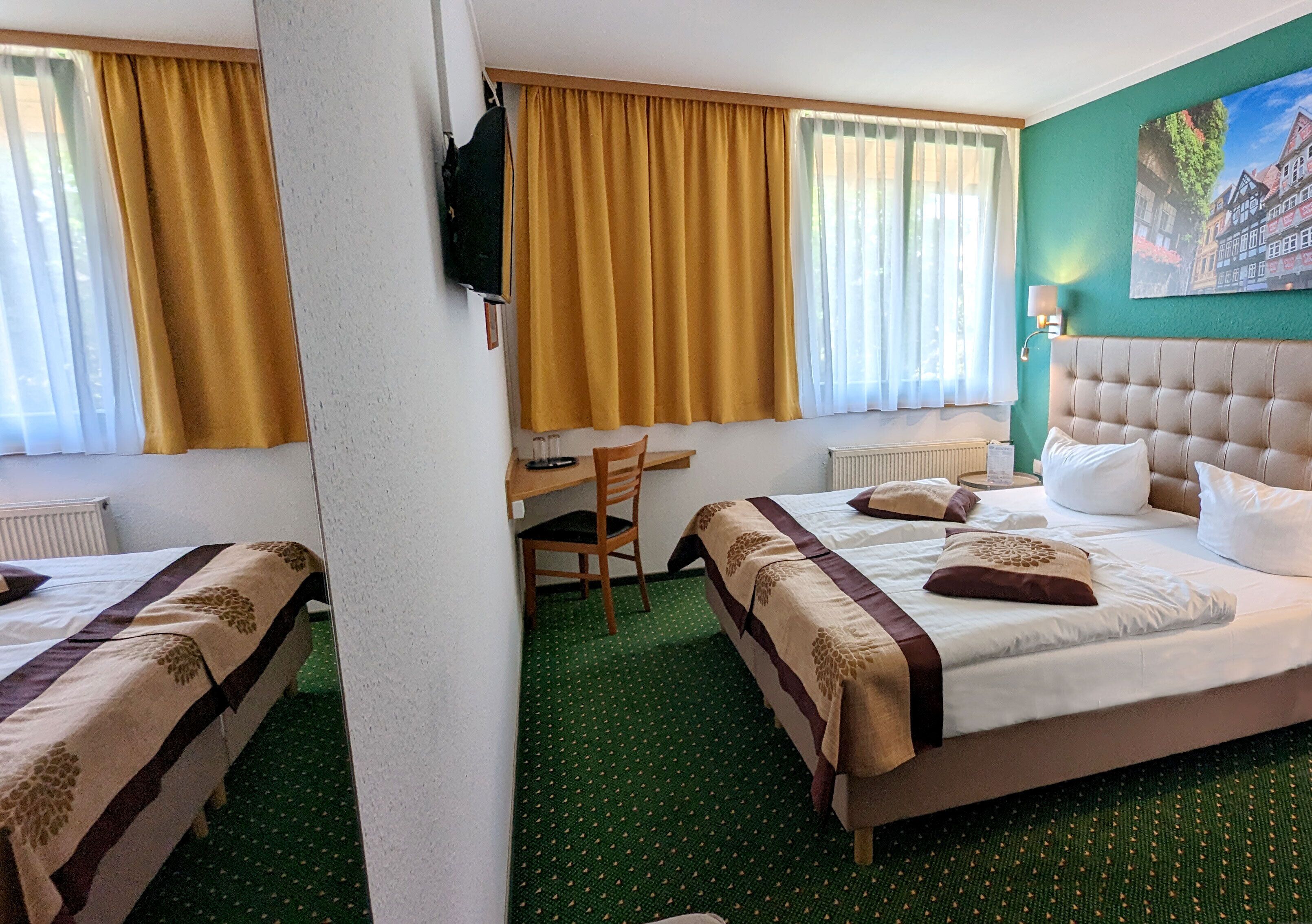 Habitación de hotel con dos camas, un escritorio, una silla, un radiador y un cuadro en la pared.