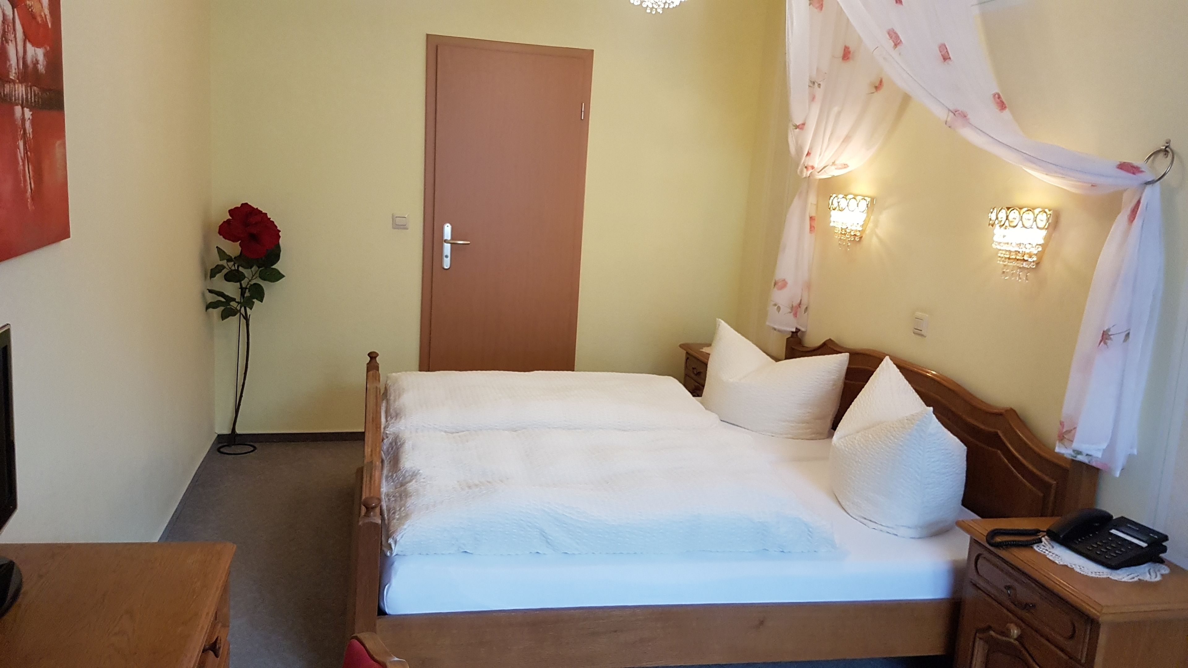 A neatly made bed with white linens and pillows, a wooden headboard, a bedside table with a telephone, and a wall-mounted light fixture.