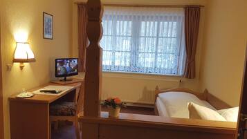 A hotel room with a wooden bed, a desk with a computer, a lamp, a window with curtains, and a radiator.