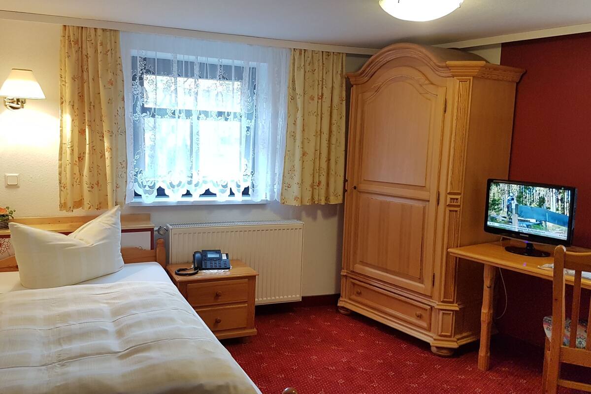 Habitación de hotel con cama, escritorio con ordenador, armario, ventana con cortinas y radiador.