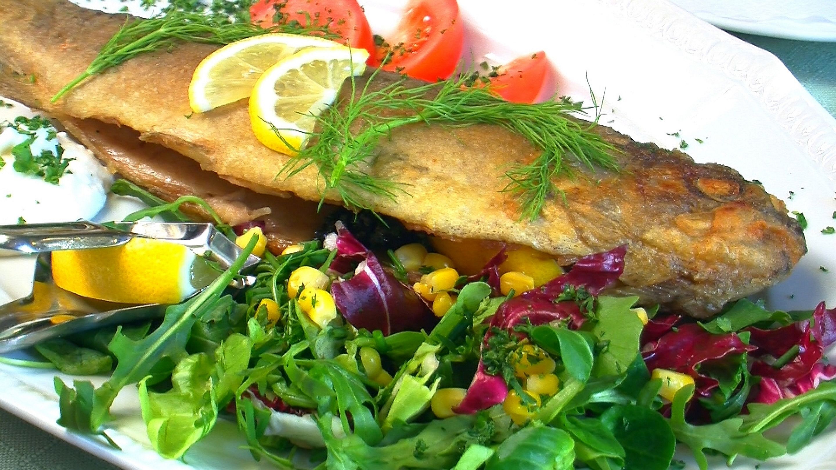 Un plato con pescado frito, rodajas de limón, eneldo y una ensalada.