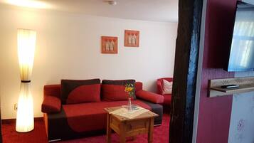 Una sala de estar con un sofá rojo, una pequeña mesita de madera con un jarrón de flores y dos cuadros colgados en la pared.