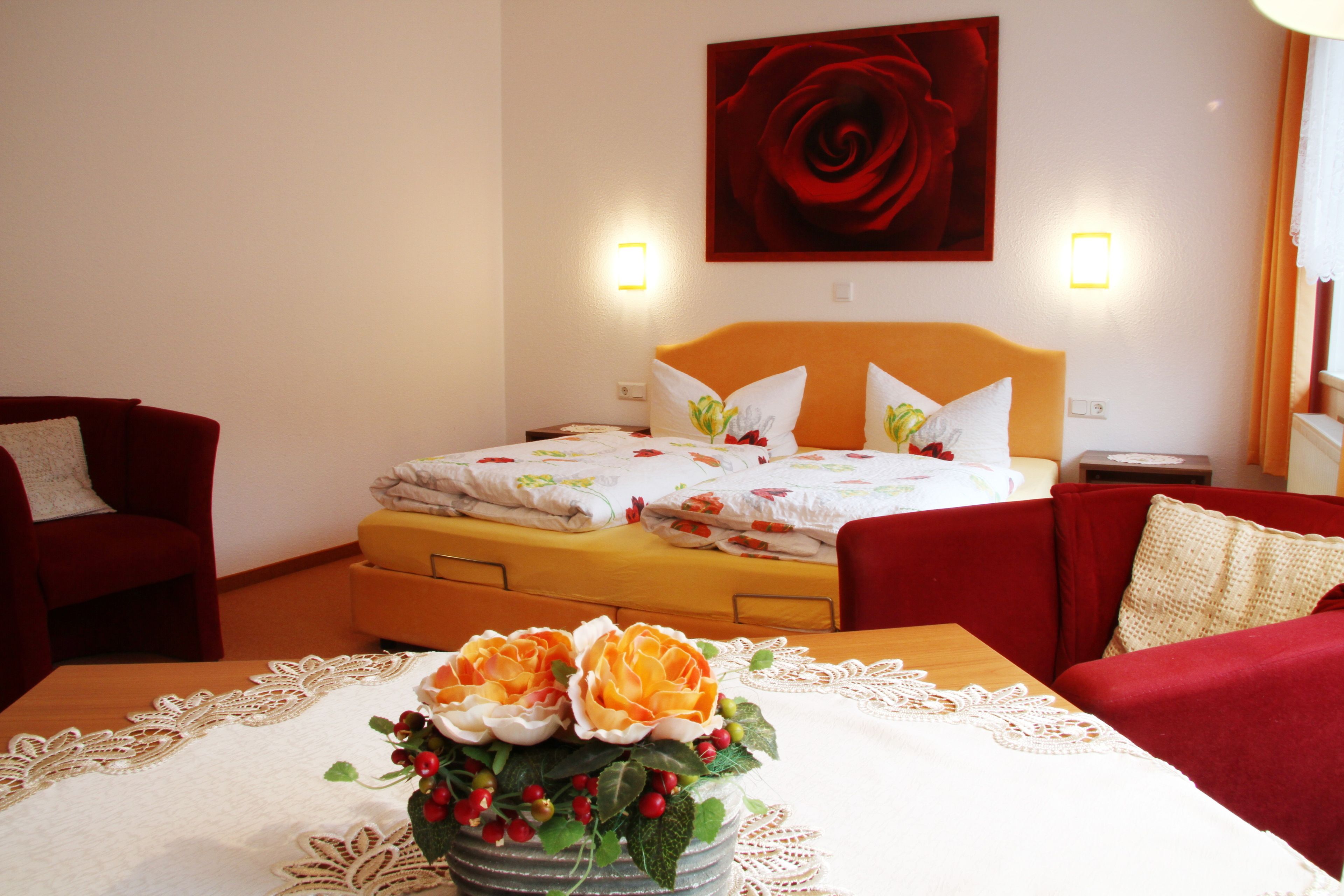 A hotel room with a bed, two red armchairs, a wooden table with a lace tablecloth, and a vase with flowers.