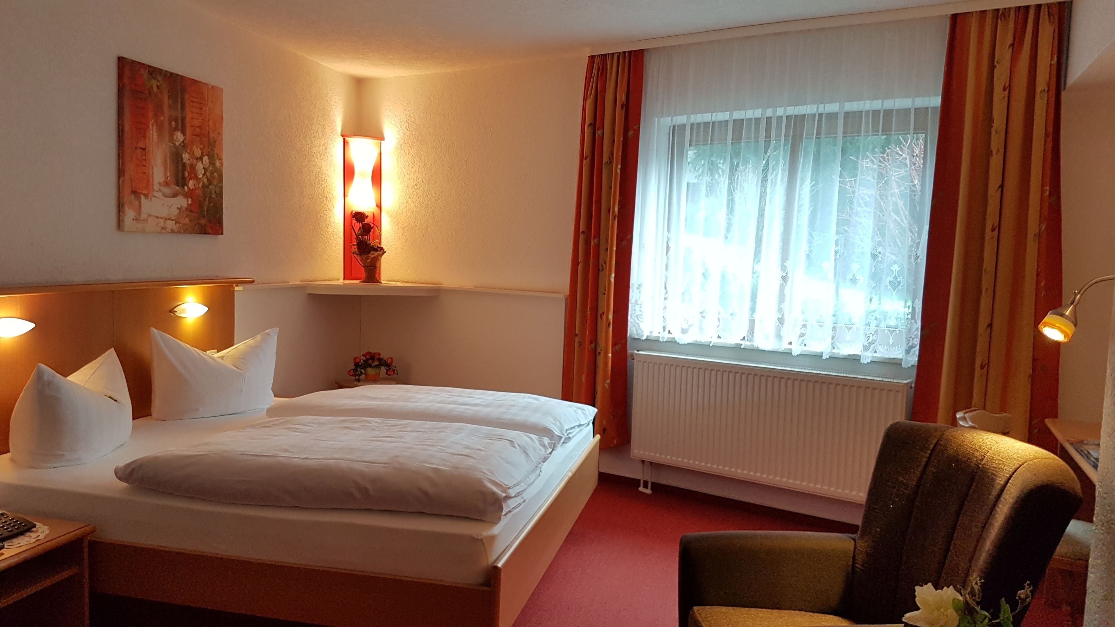 Hotellihuone, jossa on sänky, tuoli, yöpöytä, lamppu, ikkuna verhoilla sekä seinälle kiinnitetty hylly, jonka päällä on maljakko.