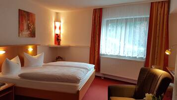 Hotellihuone, jossa on sänky, tuoli, yöpöytä, lamppu, ikkuna verhoilla sekä seinälle kiinnitetty hylly, jonka päällä on maljakko.