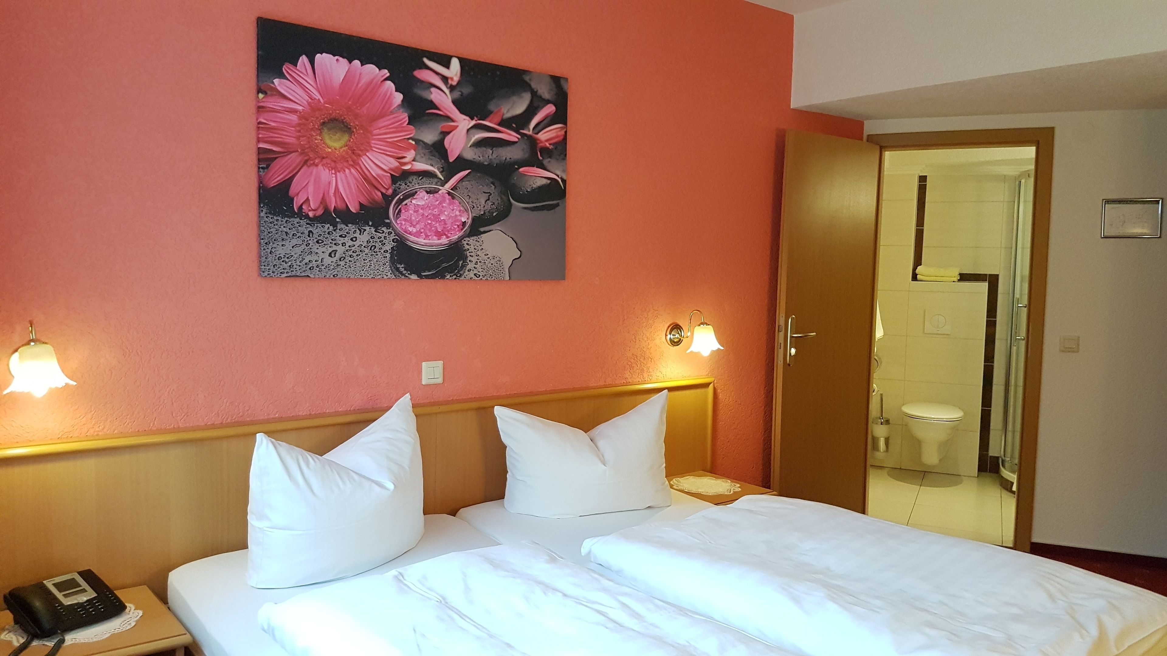 Ein Hotelzimmer mit Bett, zwei weißen Kissen, einem Nachttisch mit Telefon und einem Blumenbild an der Wand. Ein Badezimmer ist durch eine offene Tür sichtbar.