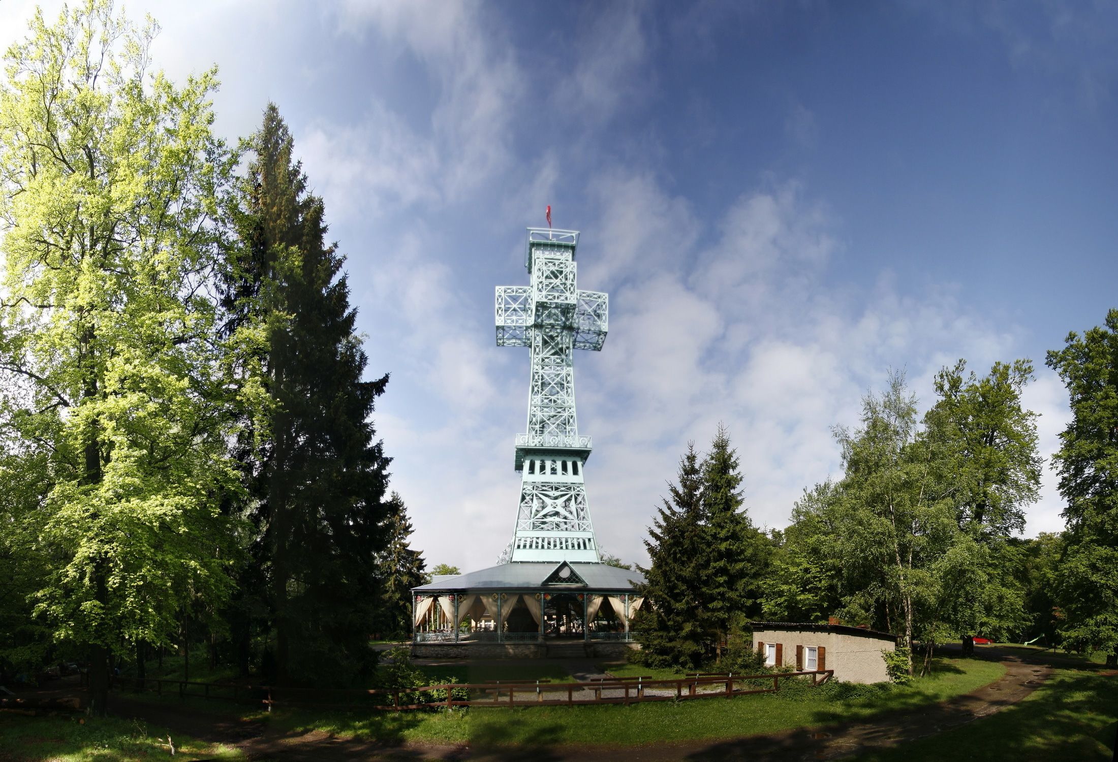 Una torre metálica alta con una cruz en la parte superior, rodeada de árboles y un pequeño edificio en su base.