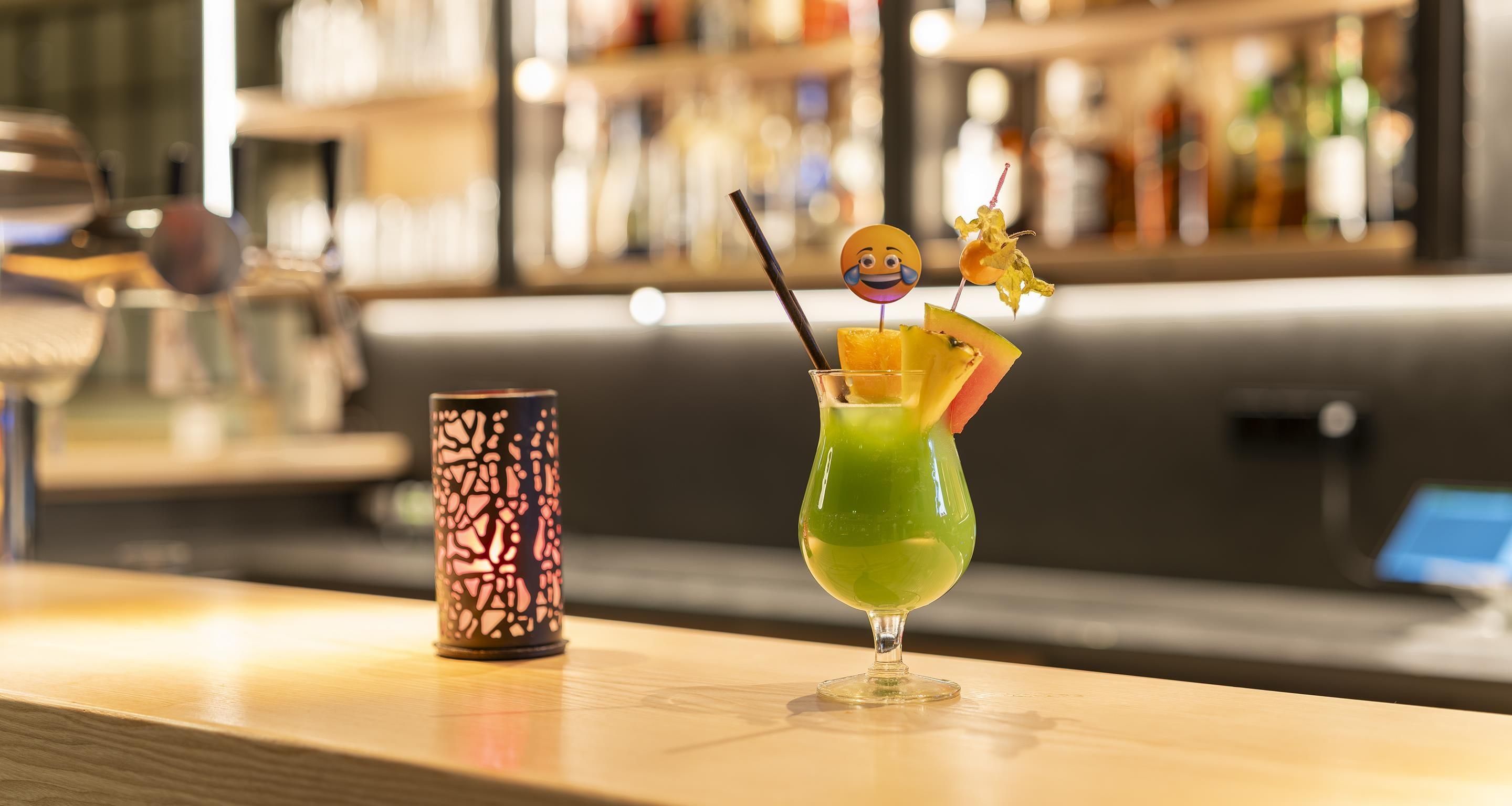 A bar setting with a cocktail glass garnished with fruit and a straw, and a decorative candle holder beside it.