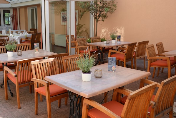 Een buitenterras met houten tafels en stoelen, plantenbakken en een menukaartstandaard.