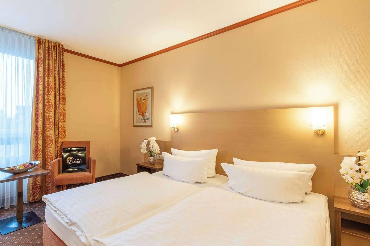 Een hotelkamer met een groot bed, een stoel, een klein tafeltje met een fruitschaal en een nachtkastje met een bloevas.