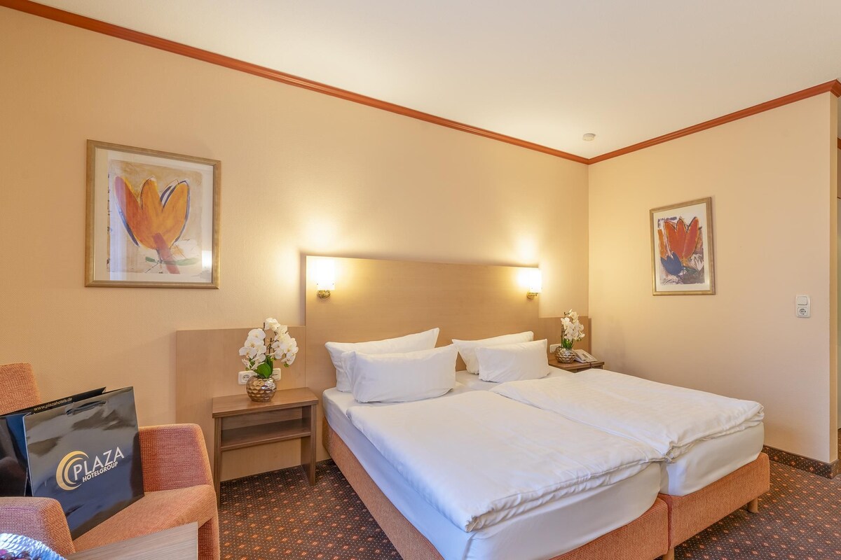 A hotel room with a double bed, a nightstand, a lamp, a framed picture on the wall, a small table with a vase of flowers, and a bag on the chair.