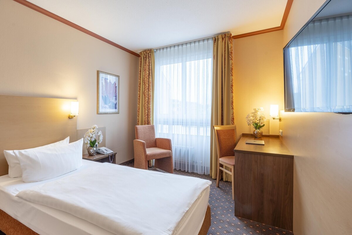 A hotel room with a bed, a desk, a chair, a nightstand, a lamp, and a window with curtains.