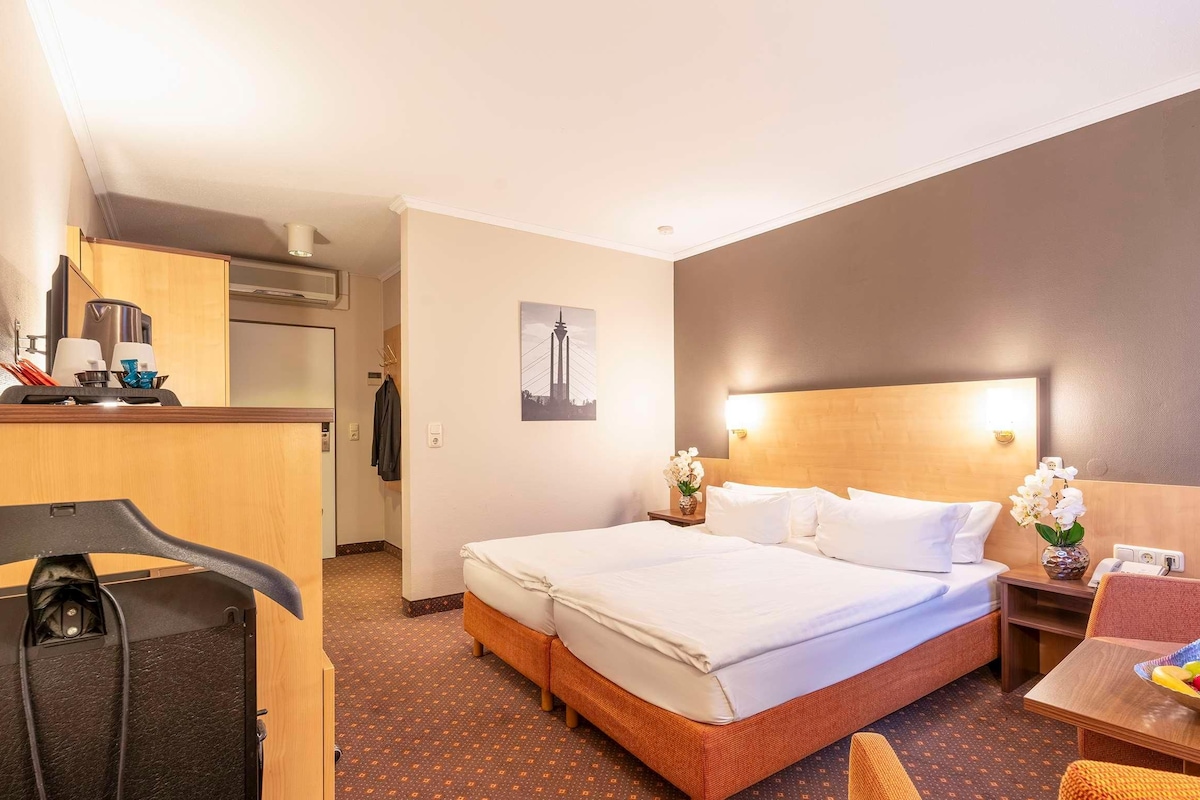 Une chambre d’hôtel avec un lit, une petite table sur laquelle se trouve un panier de fruits, une chaise, un bureau avec une lampe et un tableau accroché au mur.