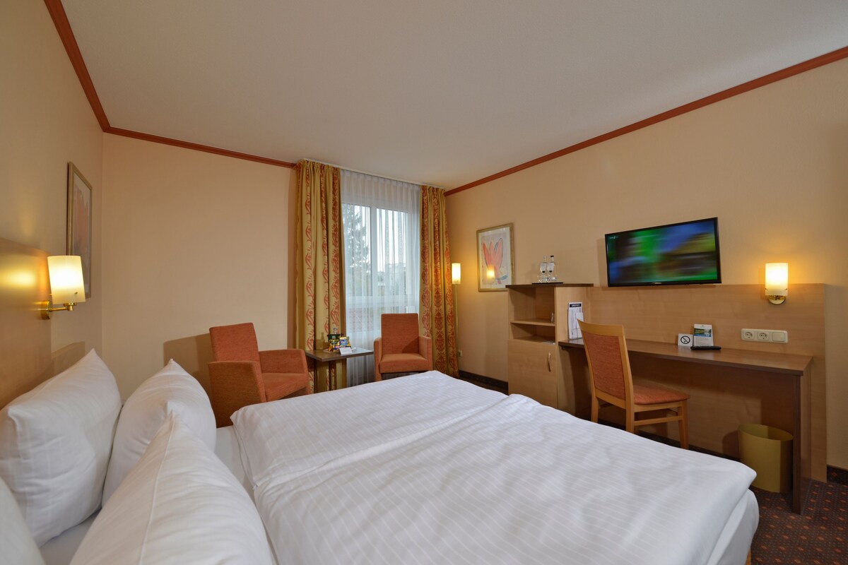 Une chambre d’hôtel avec un grand lit, deux fauteuils, un bureau, une télévision et une fenêtre avec des rideaux.