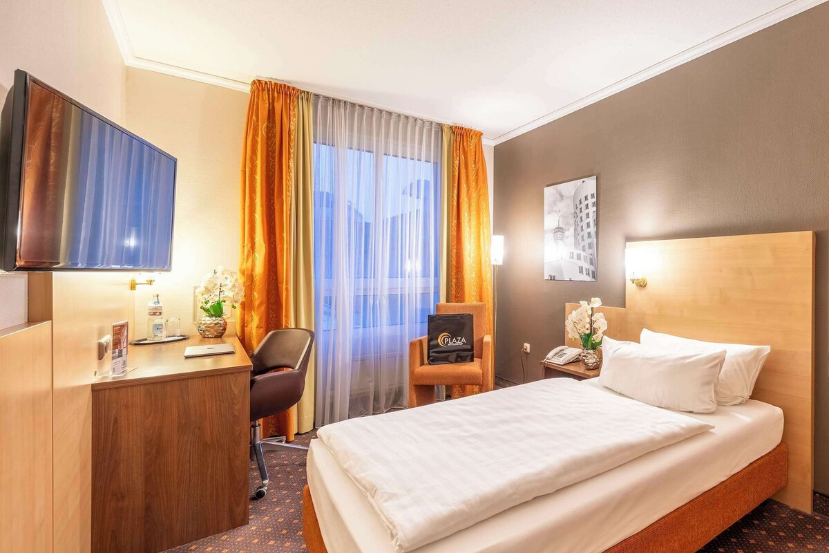 Ein Hotelzimmer mit Bett, Schreibtisch, Stuhl, Fernseher, Nachttisch und einem Fenster mit Vorhängen.